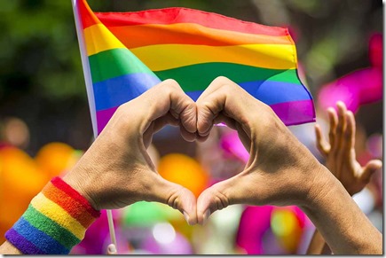 Supporting hands make heart sign and wave in front of a rainbow flag flying on the sidelines of a summer gay pride parade