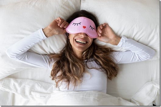 Happy millennial woman lying in bed with sleeping mask.