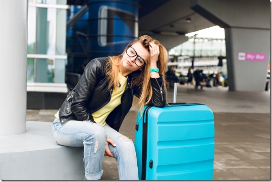 Portrait of pretty girl with long hair in glasses sitting outside in airport. She wears yellow sweater with black jacket and jeans. She leaned to the suitcase and is bored at waiting