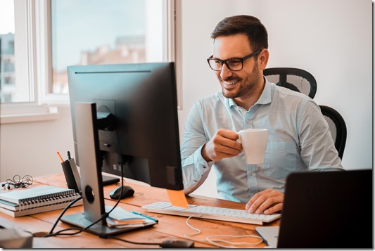 Handsome man drinking coffee while looking at computer monitor at home office.