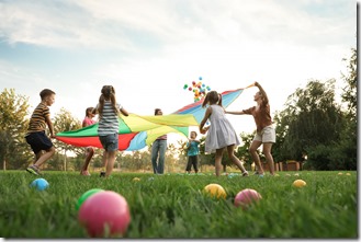 Group of children and teachers playing with rainbow playground parachute on green grass, low angle view. Summer camp activity