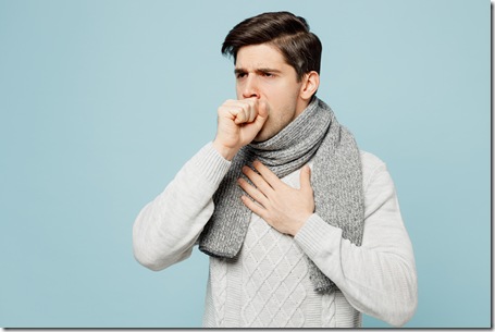 Young ill sick man wear gray sweater scarf cough sneeze cover mouth with hand isolated on plain blue background studio portrait. Healthy lifestyle disease virus treatment cold season recovery concept.